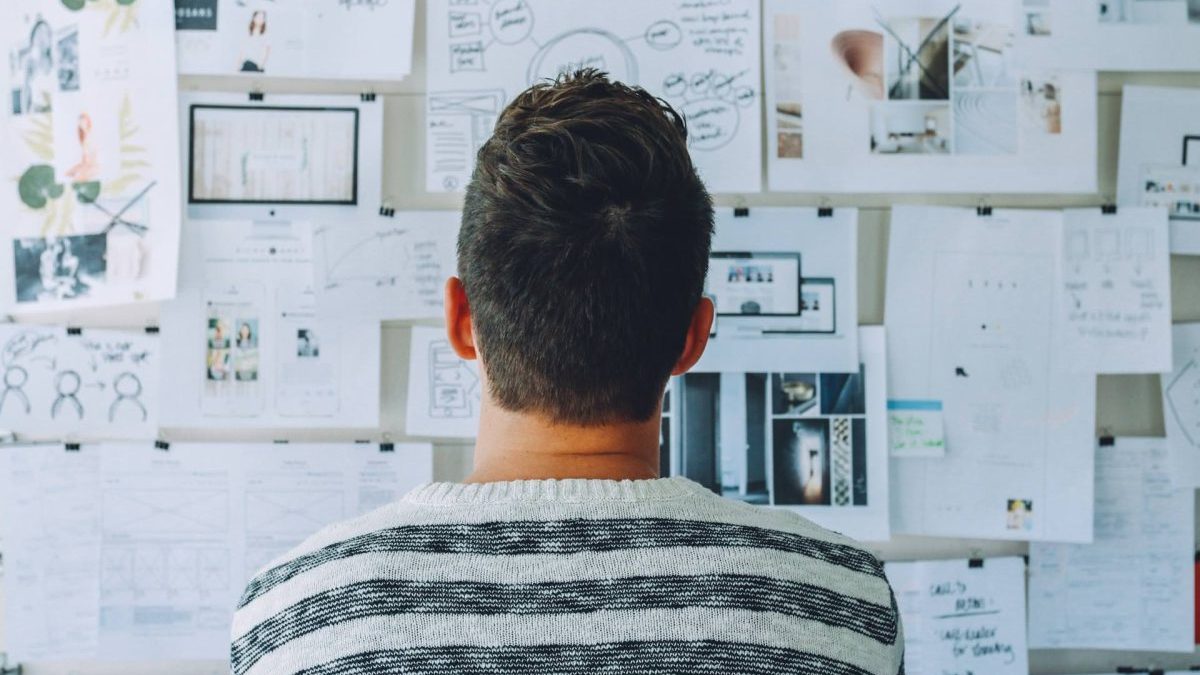 A person with short brown hair looks at a wall covered with notes, sketches, and photos, appearing to analyze or plan a project.
