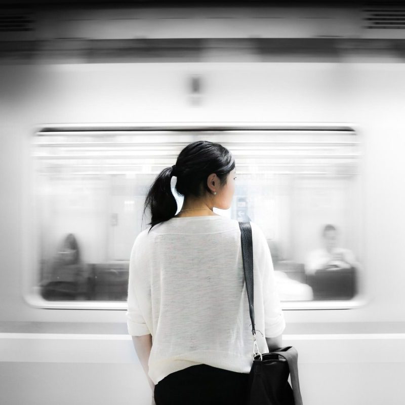 A woman with a ponytail stands on a subway platform facing a speeding train, with blurred passengers visible through the window.
