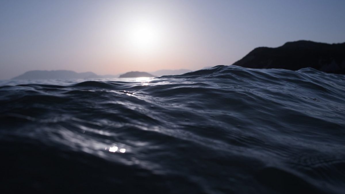 Close-up view of ocean waves with the sun low in the sky and distant hills silhouetted on the horizon.