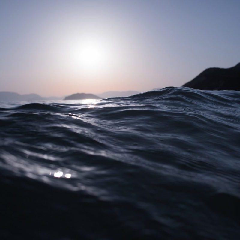 Close-up view of ocean waves with the sun low in the sky and distant hills silhouetted on the horizon.