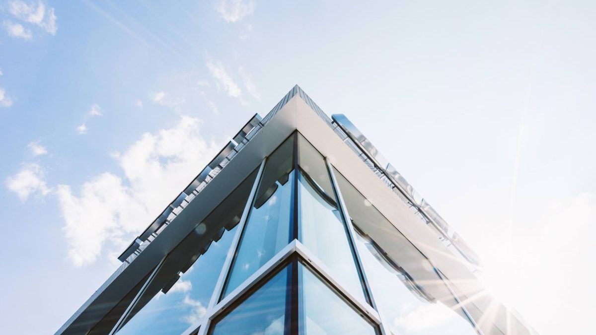 Modern glass building viewed from below with blue sky and sunlight in the background.