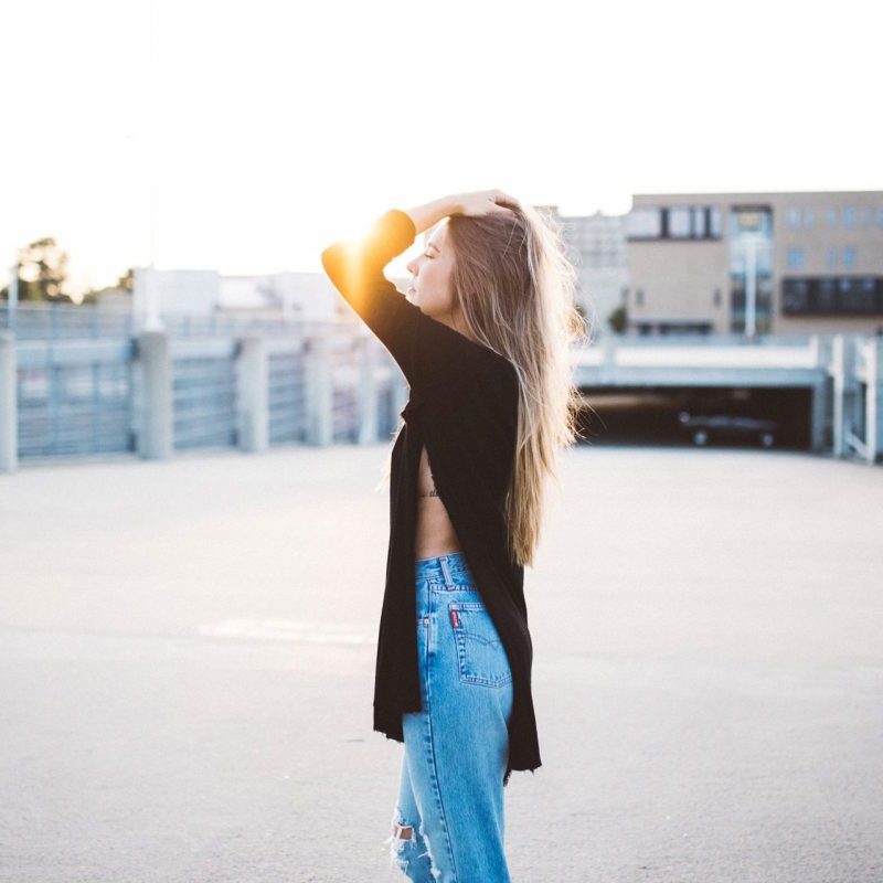 A woman stands in a parking lot at sunset, wearing a black top and blue jeans, with one hand in her hair and eyes closed.