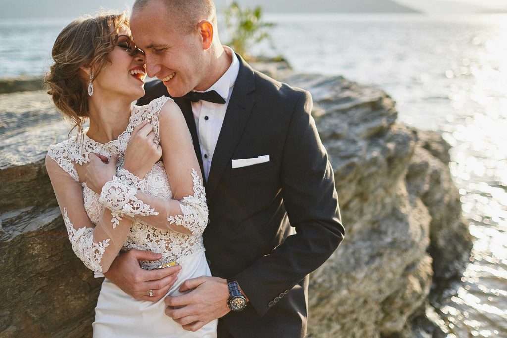 A bride and groom in formal attire smile and embrace on a rocky shoreline with water in the background.