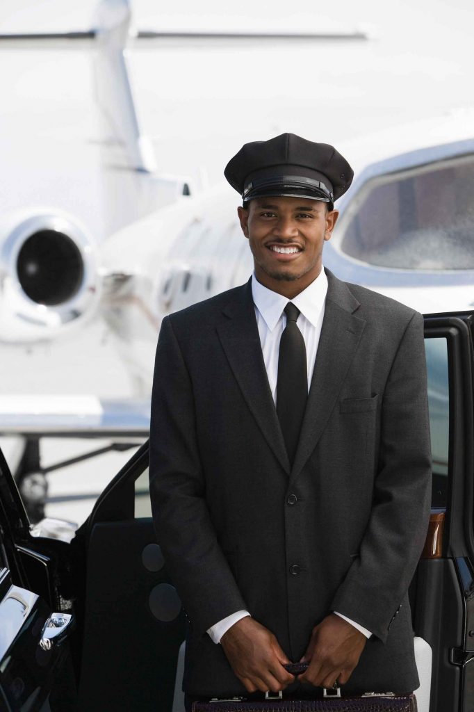 A man in a chauffeur uniform stands by an open car door with a private jet in the background.