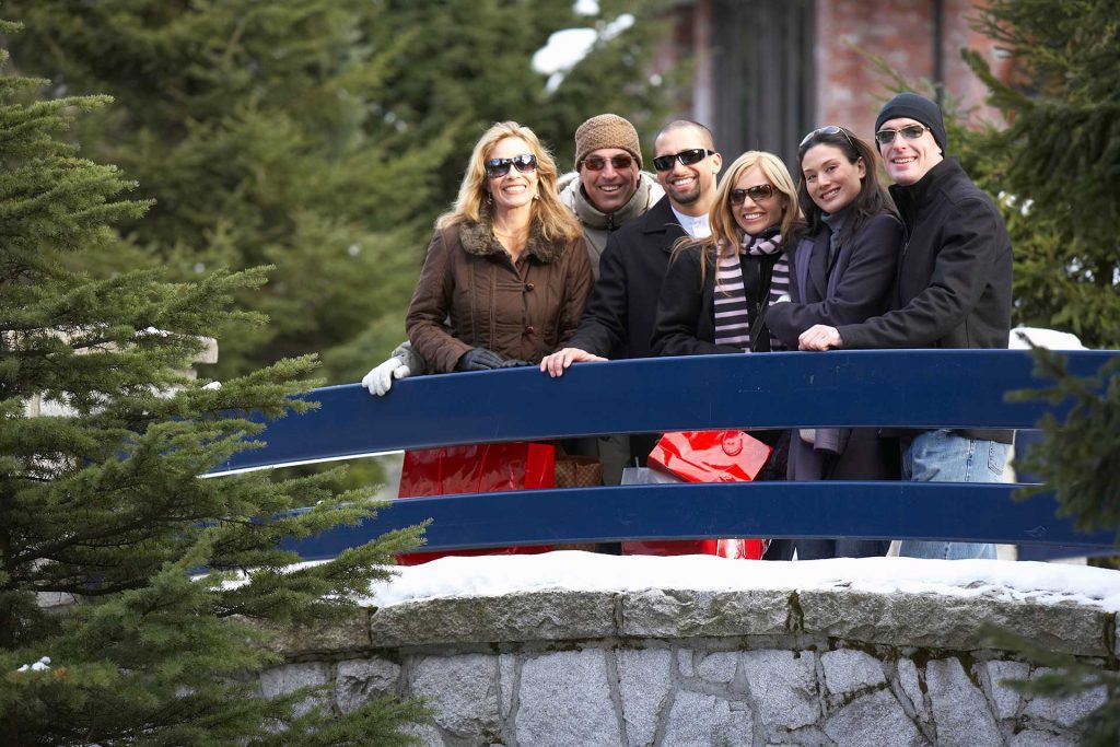 Six adults in winter clothing stand together on a blue bridge outdoors, surrounded by snow and evergreen trees.