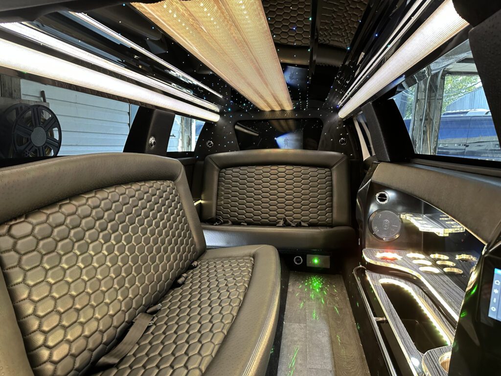 Luxurious limousine interior featuring black hexagonal-patterned leather seats, LED lighting on the ceiling and floor, and glossy surfaces with built-in controls and cup holders.