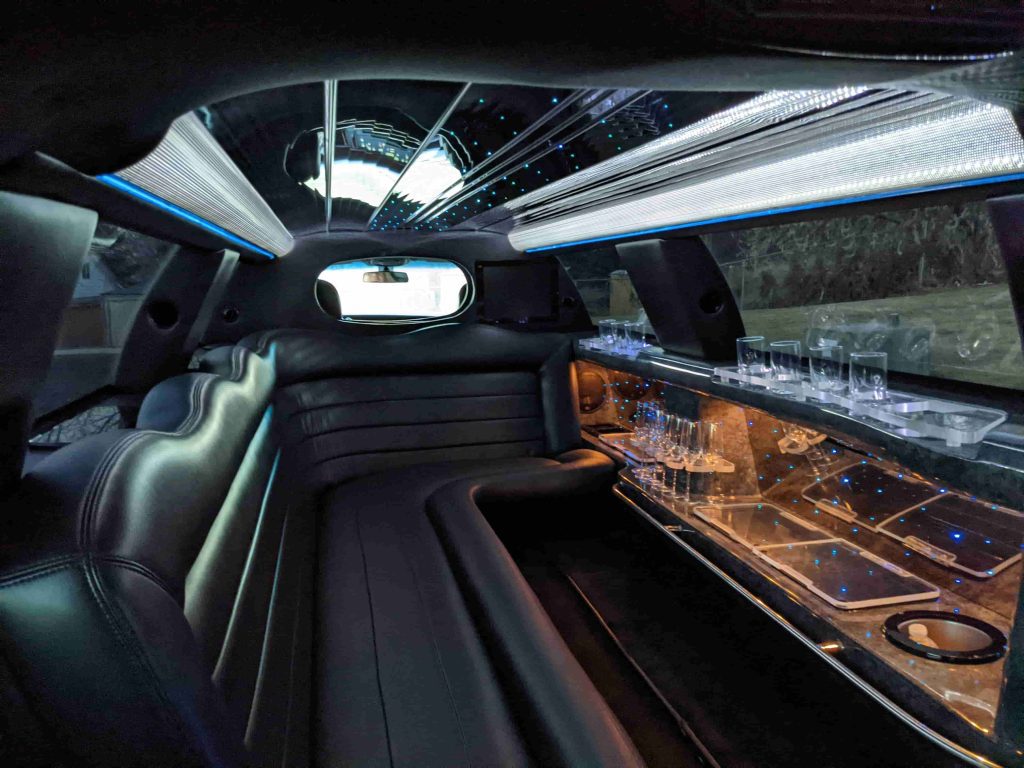 Interior of a luxury limousine showcases plush leather seats, a small bar area with glasses, mirrored surfaces, and ambient ceiling lights—just some of the refined amenities you can expect inside.