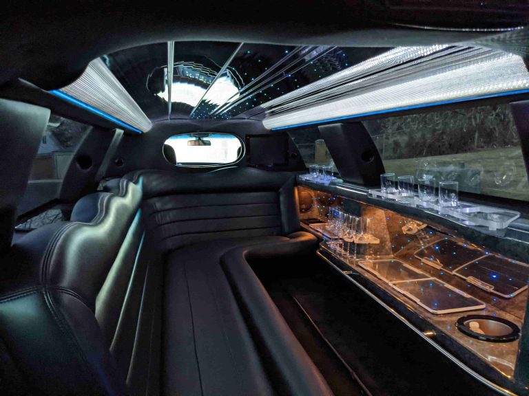 Interior of a luxury limousine showcases plush leather seats, a small bar area with glasses, mirrored surfaces, and ambient ceiling lights—just some of the refined amenities you can expect inside.