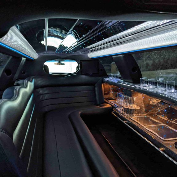Interior of a luxury limousine showcases plush leather seats, a small bar area with glasses, mirrored surfaces, and ambient ceiling lights—just some of the refined amenities you can expect inside.