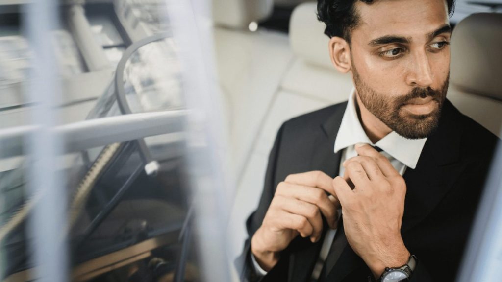 A man in a suit sits in the back seat of a car, adjusting his tie and looking out the window with a serious expression, contemplating how do I book a professional chauffeur service in Vancouver or Whistler?