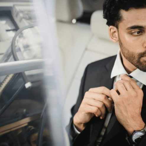 A man in a suit sits in the back seat of a car, adjusting his tie and looking out the window with a serious expression, contemplating how do I book a professional chauffeur service in Vancouver or Whistler?