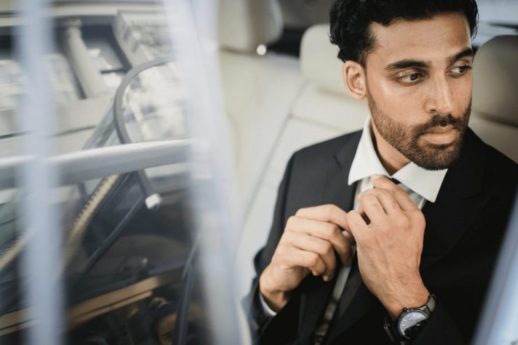 A man in a suit sits in the back seat of a car, adjusting his tie and looking out the window with a serious expression, contemplating how do I book a professional chauffeur service in Vancouver or Whistler?
