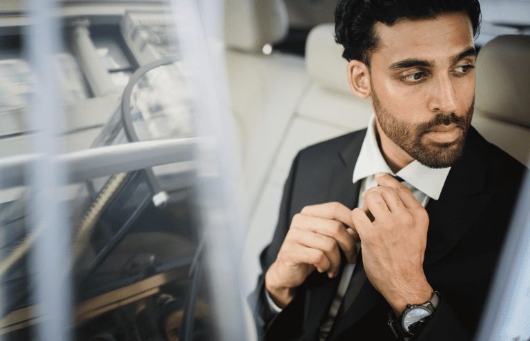 A man in a suit sits in the back seat of a car, adjusting his tie and looking out the window with a serious expression, contemplating how do I book a professional chauffeur service in Vancouver or Whistler?