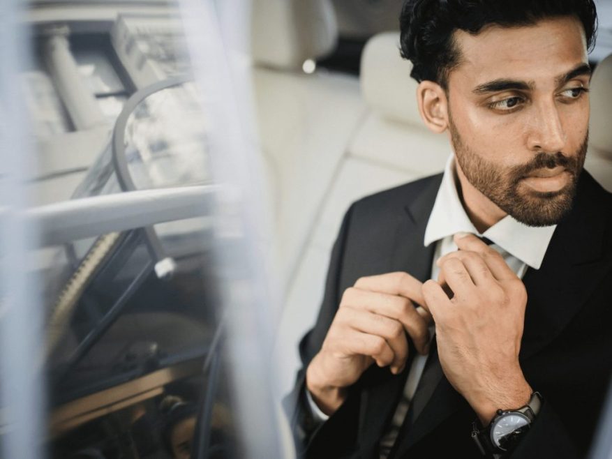 A man in a suit sits in the back seat of a car, adjusting his tie and looking out the window with a serious expression, contemplating how do I book a professional chauffeur service in Vancouver or Whistler?